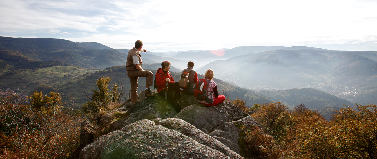 Eine Gruppe von 4 Wanderern sieht auf das Panorama des Nationalparks Nordschwarzwalds.  Im mittleren Vordergrund sind Felsen zu sehen, auf denen drei Mitglieder der Gruppe sitzen. Das linke Gruppenmitglied ist in einem Kurzarmshirt und einer Camelfarbenen sportlichen Weste Hosekombination. Er zeigt auf einen Punkt am Horizont.