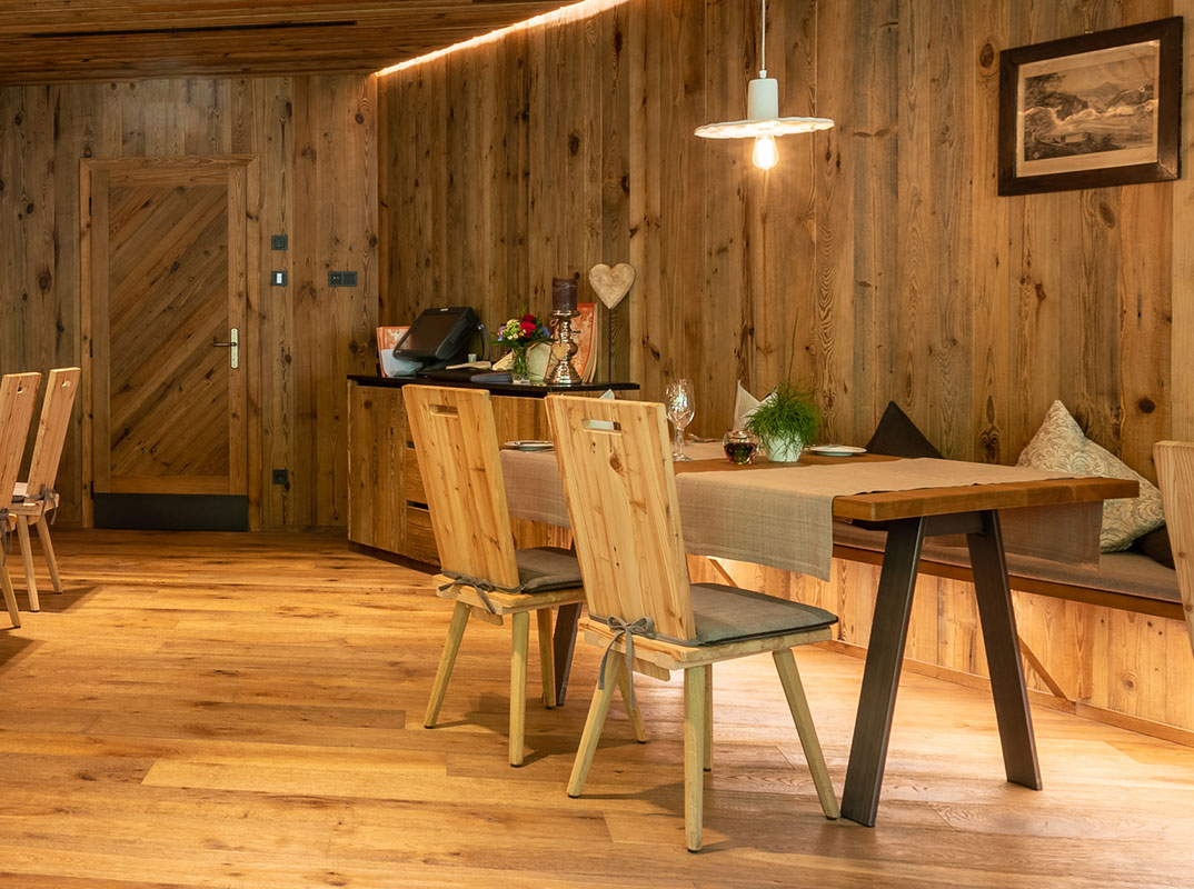 Ein Tisch in der Murgstube. Es ist ein Raum zu erkennen welcher komplett aus Holz besteht, von der Decke hängt eine weiße Lampe. In der Mitte vom Bild befindet sich ein Hölzerner Tisch. Vor den Tischen stehen 2 hölzerner Stühle und hinter den Tischen befindet sich in der Wand integriert eine hölzernere Bank. Auf dem Tische befinden sich zwei Tischdecken, diese liegen längs zu den Stühlen. Zudem stehen auf dem Tisch Gläser, graue Servietten in der Mitte des Platzes, Brotteller mit Brotmesser auf der linken Seite des Platzes, ein kleiner Blumentopf in der Mitte des Tisches und dazu eine Kerze.