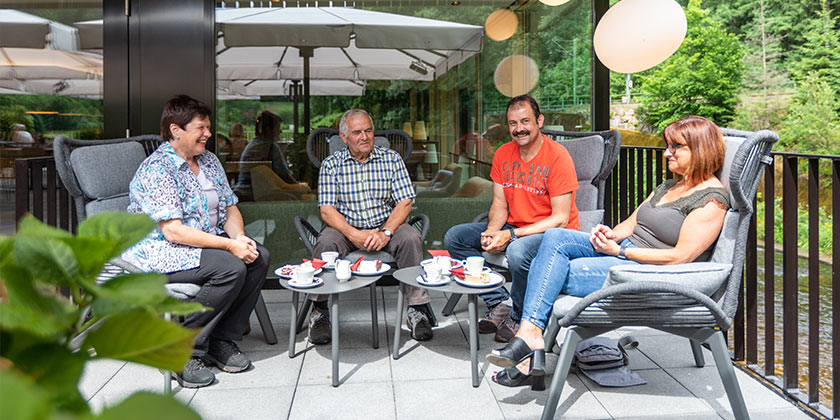 Bild von einer Vierergruppe auf der Murgterrasse. Die Gruppe besteht aus zwei Damen und zwei Herren. Diese sitzen an der Fensterfront in den dunkelgrauen Chill-Out Sesseln. Die beiden Herren Sitzen in der Mitte der Gruppe, die beiden Frauen an den jeweiligen Seiten. Die Damen auf der linken Seite des Bild hat kurze dunkle Haare trägt eine gemusterte Bluse darunter Wien weißes Shirt und schwarz Jeans sowie Wanderschuhe. Sie lacht die Damen, die Ihr Gegenübersitzt an. Der Herr in der Mitte auf der linken Seite trägt kurze graue Haare, ein blau-grün-weiß kariertes Kurzarhemd. Der Herr daneben trägt ein orangefarbenes T-Shitr. Im rechten Hintergrund des Bildes erkennt man hinter dem Geländer die Murg. Die Vierergruppe hat sich zu Kaffee und Kuchen getroffen. Die Teller stehen auf den beiden kleinen anthrazitfarbenen Loungetischen. 