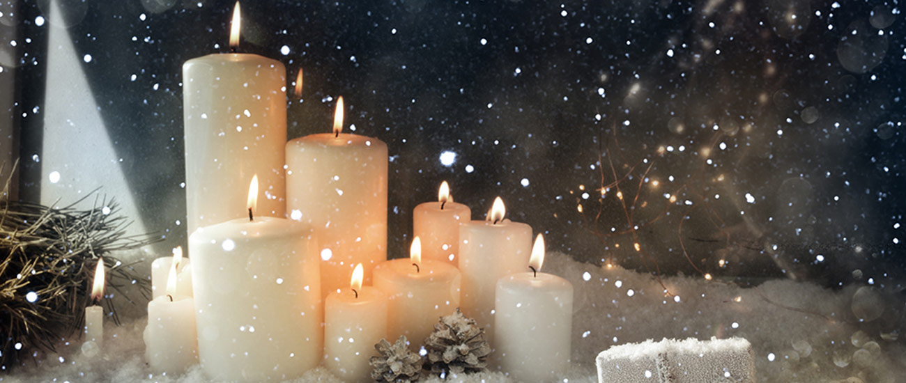 10 verschieden große brennende Kerzen stehen in einer Schneelandschaft. An den Kerzen angelehnt erkennt man zu geschneite Tannenzapfen. 