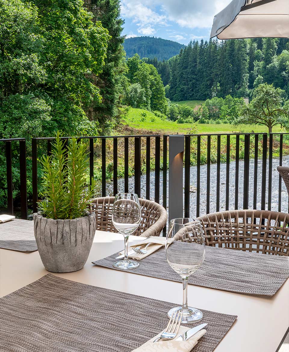 Blick von der Murgterrasse in das Murgtal. Im Vordergrund ist ein Ausschnitt des gedeckten Terrassentischs zu sehen. Zu erkennen sind insgesamt drei Gedecke. Das linke ist hierbei ausschließlich angeschnitten.  Das Gedeck besteht aus einem grauen Tisch Set. Darauf sind auf der rechten Seite des Sets Messer und Gabel in einem wachsweißen Sachen platziert. Direkt oberhalb des Bestecktes ist ein Weißweinglas platziert. Auf der rechten Seite im Eck erkennt man, dass die Sonnenschirme aufgespannt sind. 