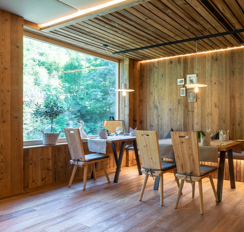 Murgstube mit Fensterplatz. Links im Bild ist ein großes Fenster zu erkennen, auf dem sich ein kleiner Baum in einem Holzkorb und eine weiße hölzerne Gans befindet. Der Raum besteht komplett aus Holz und es hängen weiße Lampen von der Decke hinunter. In der Mitte vom Bild befinden sich zwei hölzerner Tisch mit einem gewissen Abstand. Vor den Tischen stehen auf der linken Seite 1 hölzerner Stuhl und auf der rechten Seite 2 hölzerner Stühle und hinter den Tischen befindet sich, in der Wand integriert, eine lange hölzernere Bank. Auf den Tischen befinden sich graue Tischdecken, diese liegen längs zu den Stühlen. Zudem stehen auf jedem Tisch Wasser und Weingläser, graue Servietten in der Mitte von jedem Platz, Brotteller mit Brotmesser auf der linken Seite des jeweiligen Platzes, ein kleiner Blumentopf in der Mitte der Tische und daneben eine Kerze.