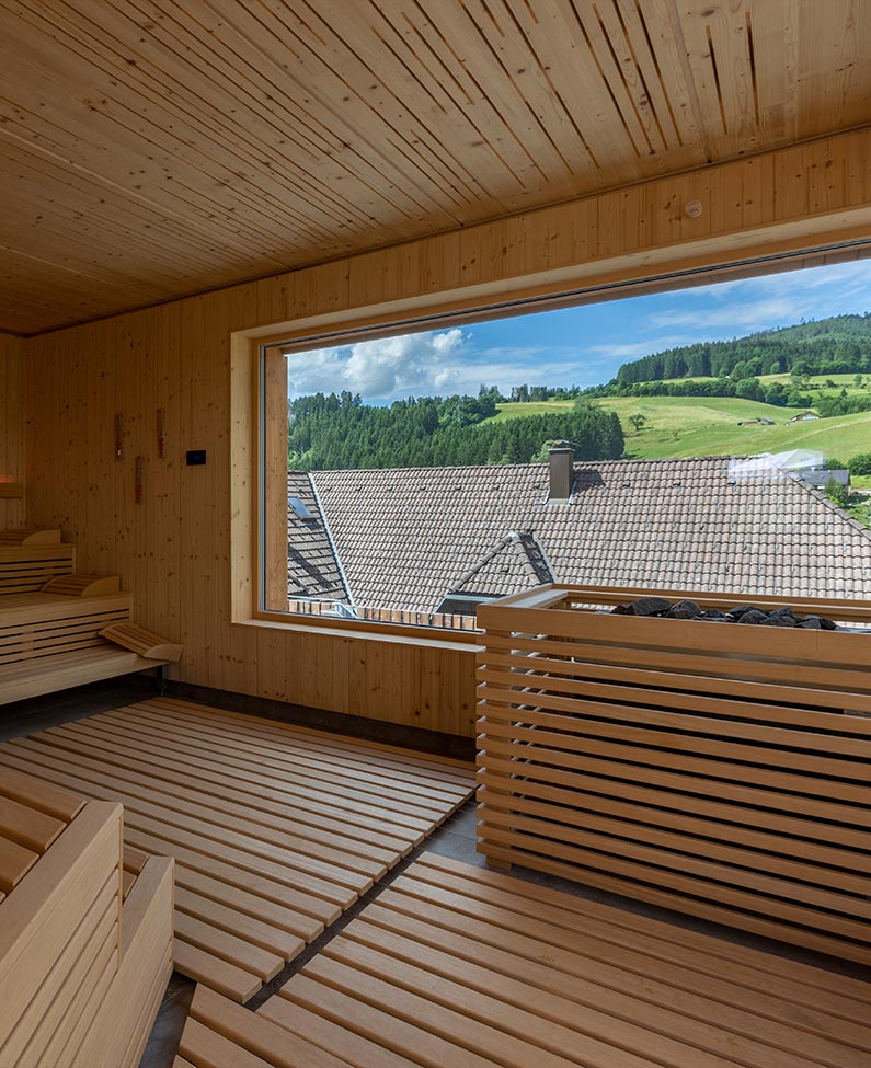 Blick aus der Holzvertäfelten Panoramasauna. Im Hintergrund ist das dunke Dach eines Hauses und die begrünten Hügel von Baiersbronn zu sehen. In der rechten Reite des Bildes ist das Aufgusselement, auch holzvertäfelt mit seinen heißen schwarzen Steinen, zu sehen.