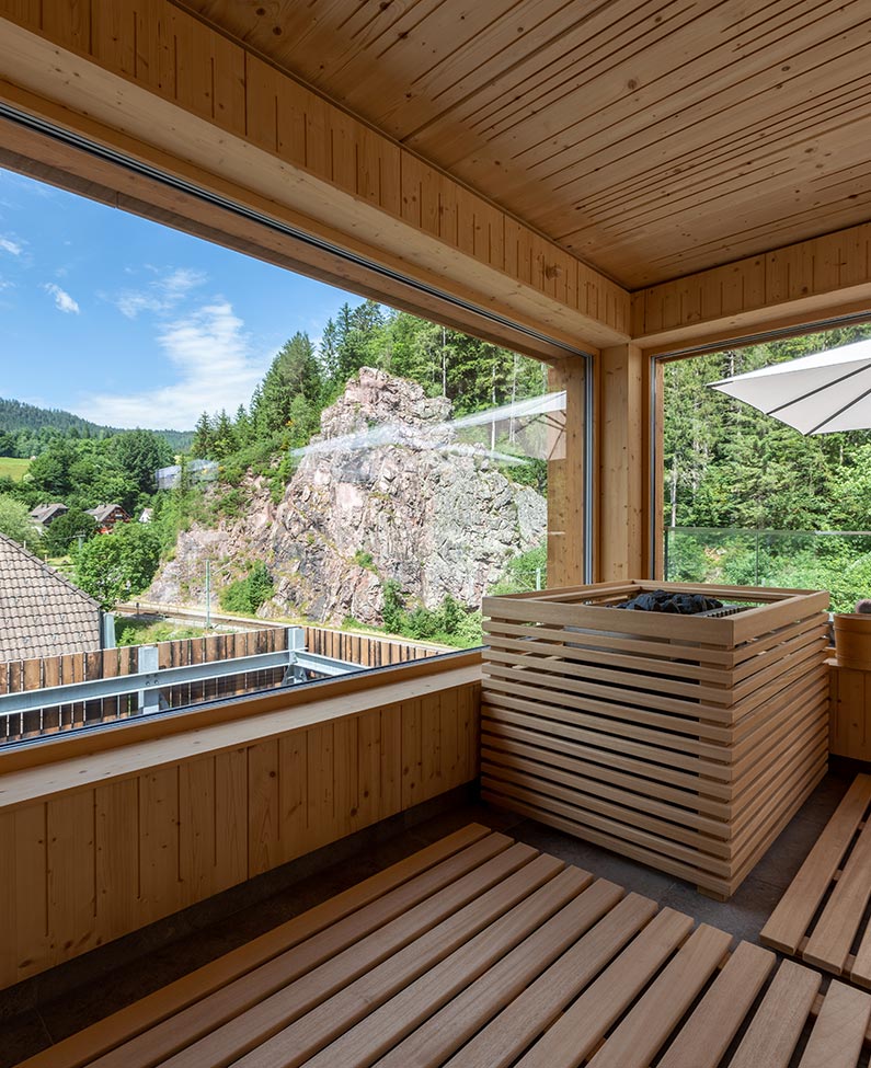 Ausblick auf den Murgfels auf einer Sauna. Im mittigen Hintergrund ist der Murgfels zu sehen. Die Sauna ist mit Panoramafenstern ausgetattet, sodass man den Blick genießen kann. Diese befinden sich an der linken Seite und im rechten Hintergrund.Vor den Fenstern steht ein mit Holz verkleideter Aufgusskessel.