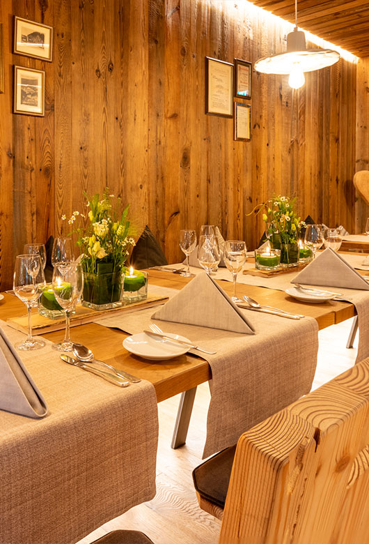 Blick auf eine Tafel in der Murgstube. Im Hintergrund ist die Wandvertäfekung zu sehen. An dieser hängen sowohl links im Eck als auch auf der rechten Seite gerahmte Bilder und Fotografieren. Auf der linken Seite sind eine Querfotografierte und eine hochformatige Fotografie aufgehängt. Auf der rechten Seite sind eine hochformatige größere Illustration und rechts daneben zwei kleinere hochformatige Bilder an der Vertäfelung angebracht. Schräg vor der rechten Wanddeko ist eine Hängelampe mit schirm zu erkennen. Das zentrale Element des Bildes bildet die gedeckte Tafel mit vier Tischläufern nebeneinander. Diese verlaufen vom Vordergrund in den Hintergrund und sind grau meliert. In der Mitte der Tafel stehen zwei Dekoelemente in Grün. Diese sind länglich und bestehen aus links und rechts zwei grünen Kerzen und in der Mitte einem kleinen Arrangement aus weißen Rosen Moosboden und Farnen.  Vor dem linken Dekolement ist gut das Gedeck des Tisches zu erkennen. Dieses besteht von rechts nach links aus einem Wasserglas, dahinter – Richtung Tischmitte – einem Weißweinglas, vor dem Wasserglas sind rechts ein Löffel und links daneben ein Messer zu sehen. Mittig ist eine dreieckig gefaltete Serviette platziert . Links sieht man eine gabeln, ein Brotteler und das Brotmesser. Die Weiteren Gedecke sind analog diesem Gedeck links und Rechts daneben sowie gegenüber platziert.