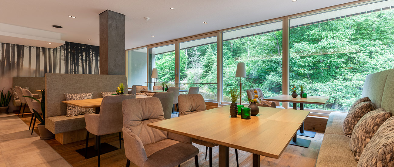 Blick auf die Sitzgelegenheiten in der Rheinholz Bar. Im Hintergrund ist die Panoramafront mit Blick auf die Murg zu sehen. Die Fensterfront gibt in dieser Perspektive den Blick auf den gegenüberliegenden Wald frei. Direkt vor dem Fenster steht ein Tisch mit Tischdekoration. Auf der linken Seite dieses Tisches steht ein grau-gepolsterter Stuhl, auf der rechten Seite ist eine raue Sitzbank mit gepolsterter Rücklehne und gepolsterter Sitzfläche integriert. Auf der Sitzbank sind drei Kissen im Rautenmuster platziert. Die Sitzbank erstreckt sich sowohl über die Tischkombi direkt am Fenster als auch bis zum Tischarrangement im Vordergrund. An das Tischarrangement auf der rechten Seiten reiht sich links gespiegelt ein weiteres Tischarrangement an.  Im Hintergrund stehen zwischen den beiden Tischen Stehlampen mit weißen Lampenschirmen. Im linken Hintergrund ist eine schwarz-weiß Tapete zu sehen, die den Schwarzwald bei herbstlicher Stimmung zeigt.