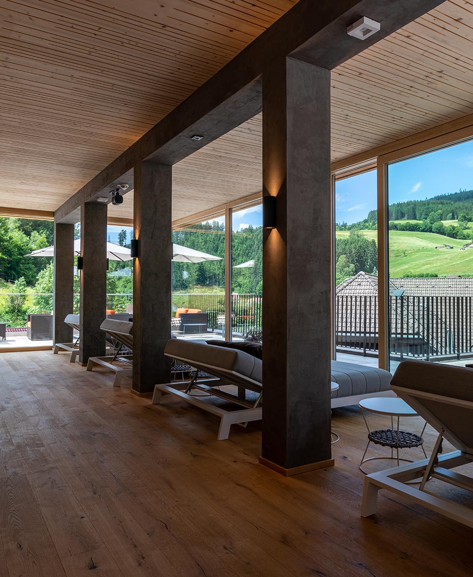 Blick zwischen die Säulen in Betonoptik auf die Daybeds im Ruheraum. Im Hintergrund ist die Glasfassade und deru Blick auf die Hügel in Baiersbronn zu sehen. Auf der linken Seite ist zudem der warme Holzboden des Ruheraums gut erkennbar.