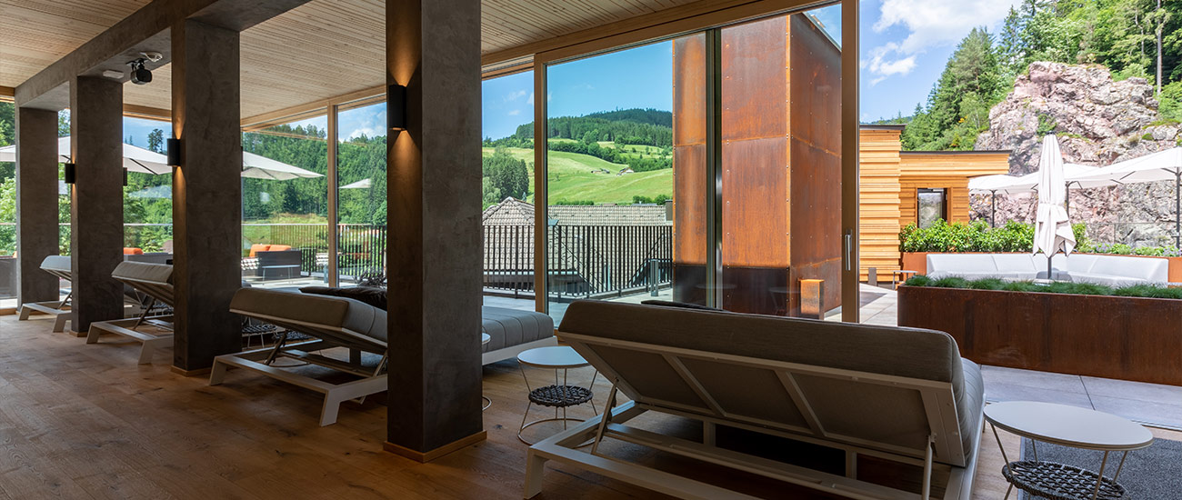 Im Vordergrund sind die Rückseiten der dunkelgrauen Daybeds des Sky Spa Ruheraums zu sehen. Diese gewähren den Blick auf die Hügel von Baiersbronn auf der linken Seite und der Terrasse auf der rechten Seite. Auf dieser sind auch drei beige Sonnenschirme aufgespannt