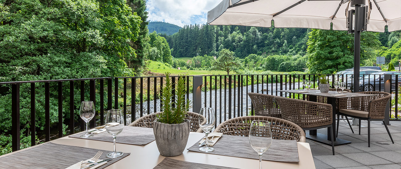 Blick von der Murgterrasse in das Murgtal. Im Vordergrund ist ein gedeckter Terrassentisch zu sehen. Dieser ist für vier Personen gedeckt. Das Gedeck besteht aus einem grauen Tisch Set. Darauf sind auf der rechten Seite des Sets Messer und Gabel in einem wachsweißen Sachen platziert. Direkt oberhalb des Bestecktes ist ein Weißweinglas platziert. Das Bild gibt den Blick auf drei der vier Gedecke frei. In der Mitte des Tischs ist eine Grünpflanze in einem dunkelgrauen Topf platziert. Auf der rechten Seite im Eck erkennt man, dass die Sonnenschirme aufgespannt sind. Das schwarze Geländer verdeckt den Parkplatz auf der rechten Seite gut.