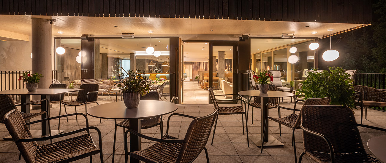 Blick von der Murgterrasse in Richtung der beleuchteten RheinHolz Bar bei Nacht. Im Hintergrund entdeckt man die Fensterfront, welche den Blick auf die hell erleuchtete RheinHolz Barfreigibt. Im Vordergrund erkennt man im dunkeln die Umrisse der Sitzgelegenheiten auf der Terrasse. Die Beleuchteten Dekolampen links und rechts vor der Fensterfront, spiegeln sich im Glas.